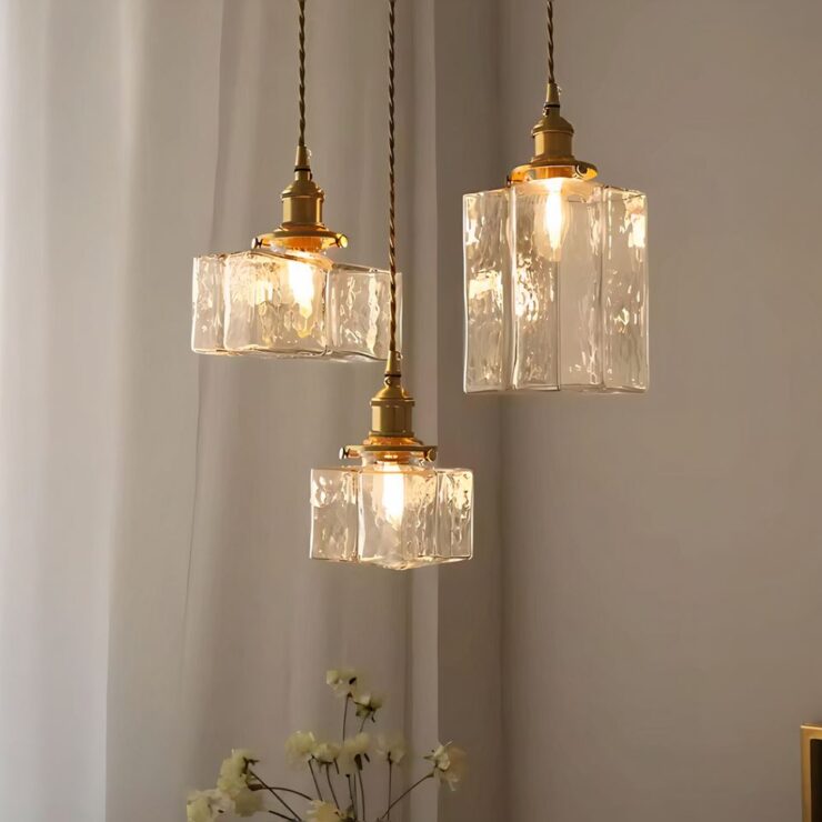 3 lustres cubiques en verre texturé avec une ampoule allumée, suspendus dans une pièce lumineuse avec des rideaux en fond et des fleurs en décoration.