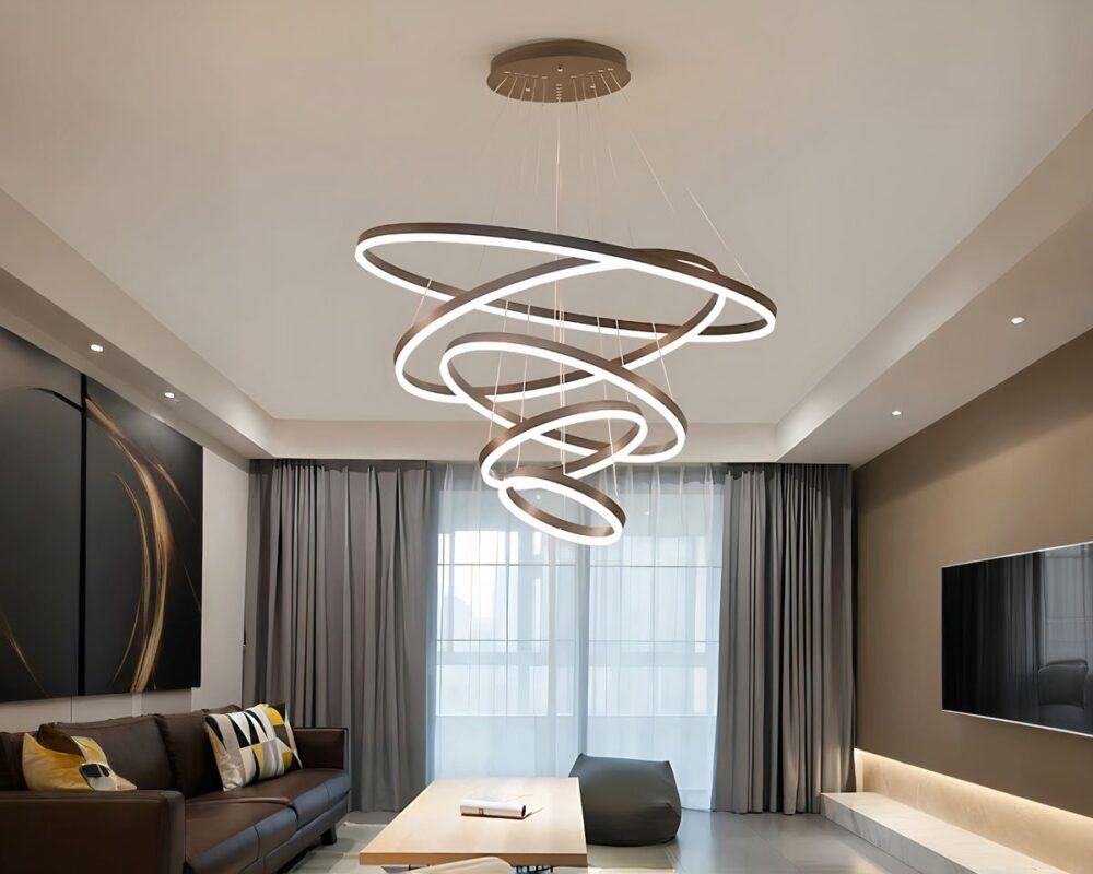 Grand lustre avec cercles suspendus en quinconce de couleur marron, le dessous étant le cercle lumineux. Dans un salon avec une grande baie vitrée en fond avec rideau gris. A gauche, un canapé style similicuir marron, sous le lustre une table basse beige. Le mur de droite est marron avec un écran plat fixé dessus.