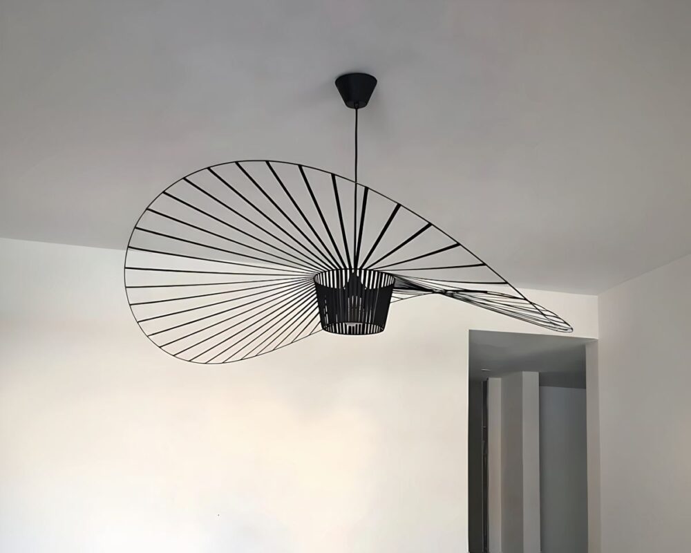 Grand lustre rétro style vertigo de couleur noire en forme de chapeau capeline à l'envers. La forme ronde est ondulée et présente des rayons ajourés. Fixé au plafond blanc avec un mur blanc au fond.
