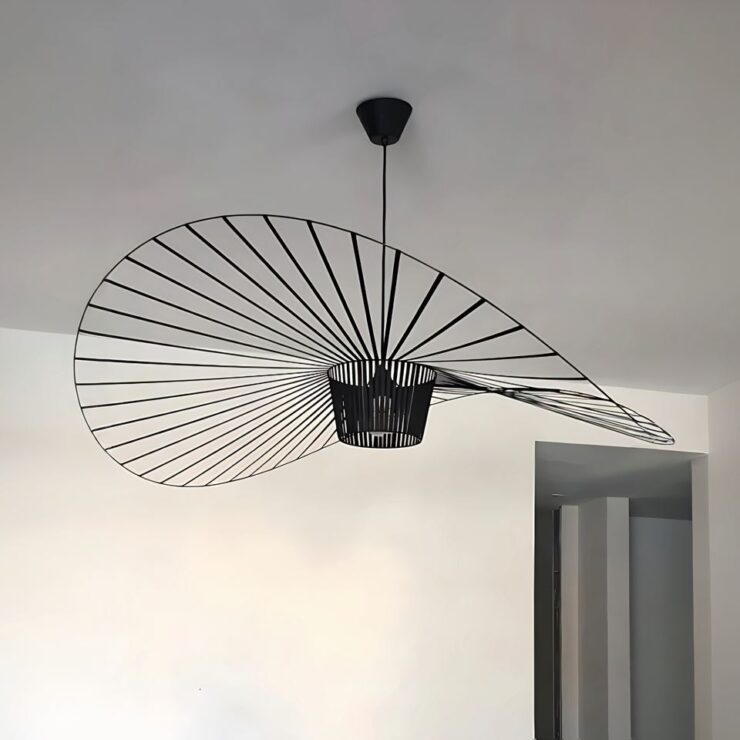 Grand lustre rétro style vertigo de couleur noire en forme de chapeau capeline à l'envers. La forme ronde est ondulée et présente des rayons ajourés. Fixé au plafond blanc avec un mur blanc au fond.