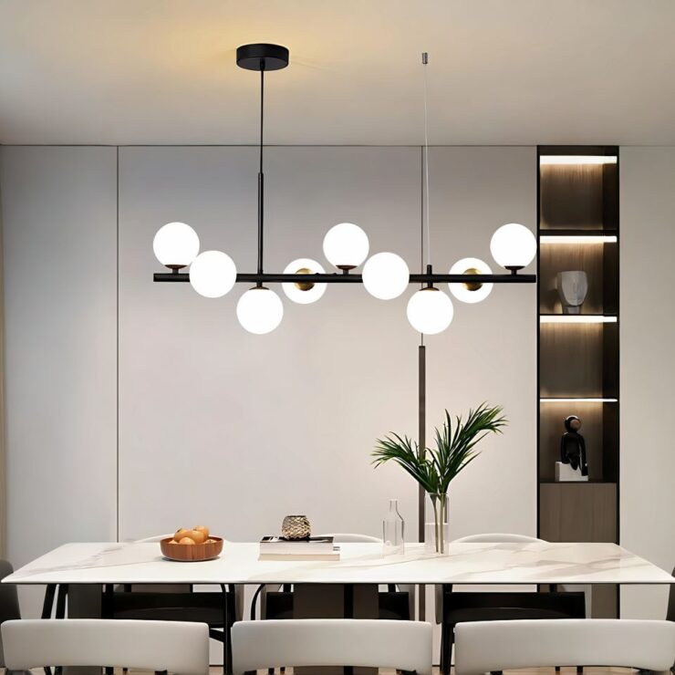 Grand lustre suspendus avec 9 boules lumineuses sur une barre horizontale noire. Le lustre est suspendu par des câbles. Il est placé au-dessus une grande table beige avec des chaises assorties sur laquelle sont posésun plat marron avec fruits et un vase transparent avec feuillage. En fond, un mur blanc avec à droite une étagère intégrée marron avec 4 cases éclairées.