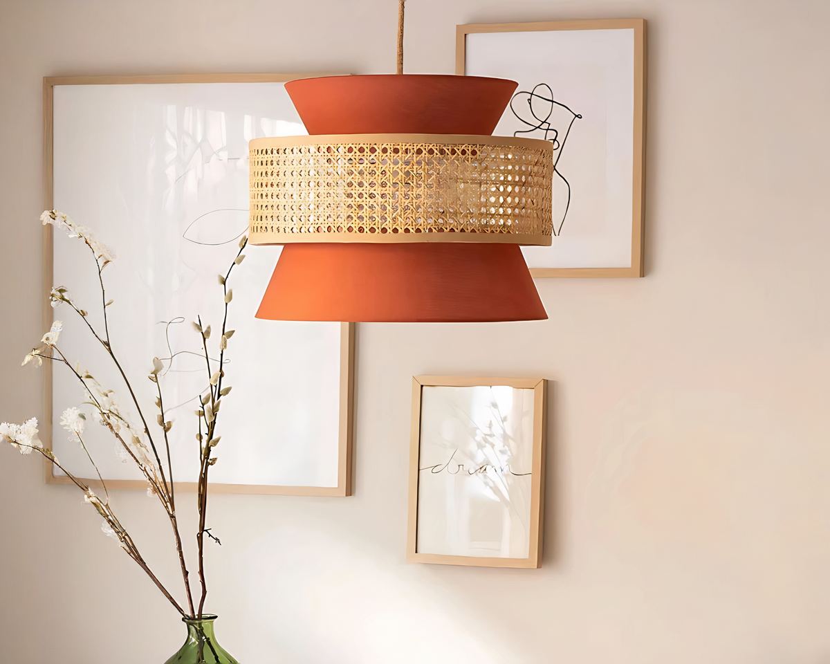 Le lustre de bureau en bambou et tissus orange sur un mur du fond beige. Des cadres blanc minimaliste y sont accrochés et on peut apercevoir des branches de petites fleurs séchées avec le goulot très certainement d'une Dame Jeanne.