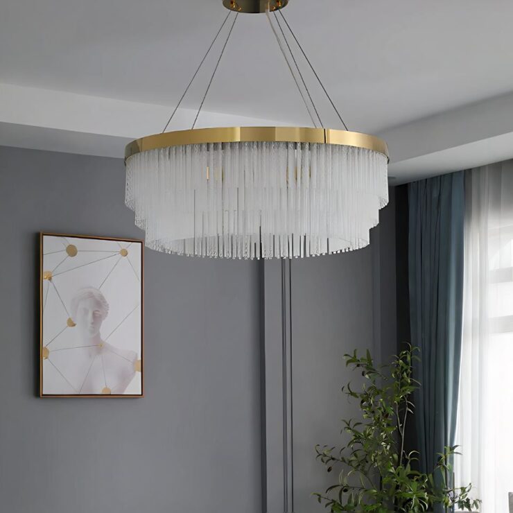 Lustre doré à pampilles en forme de bâtons en verre. Le lustre rond est suspendu par 4 câbles. Le socle au plafond et la base du cercle sont dorés. Les murs de la pièce sont gris. Un tableau à gauche et une fenêtre à droite.