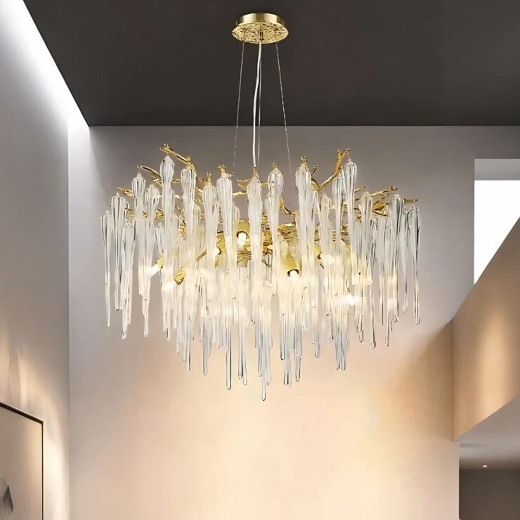Lustre doré en forme de branches avec grandes pampilles style cristal. Le lustre est suspendu à un plafond gris. Les ampoules sont allumées. Les murs autour sont blancs et éclairés.