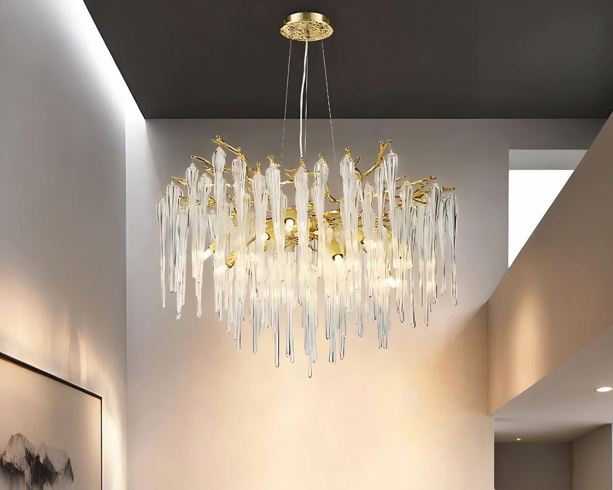 Lustre doré en forme de branches avec grandes pampilles style cristal. Le lustre est suspendu à un plafond gris. Les ampoules sont allumées. Les murs autour sont blancs et éclairés.