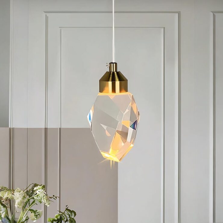 Lustre LED cristal suspendu à plusieurs facettes laissant passer la lumière. Il est suspendu par un câble blanc, le cache de la douille est doré. En fond un mur blanc et beige avec une plante verte en bas à gauche.