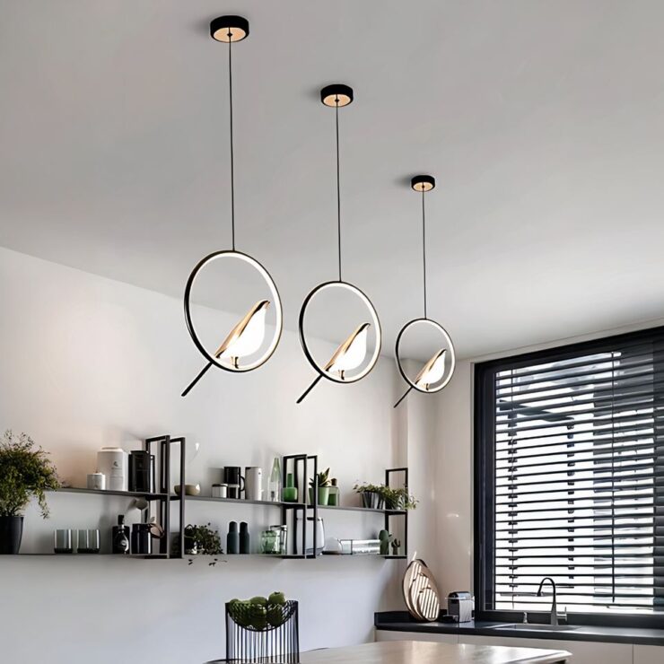 Nous sommes dans une cuisine ouverte avec trois lustres LED suspendus avec oiseau perché sur une cercle noir avec l'intérieur lumineux. Le cercle est suspendu à un câble noir qui lui même est tenu par un socle rond noir fixé au plafond. A droite on voit une fenêtre avec un encadrement noir et des persiennes noires au-dessus un évier sur un plan de travail noir avec des placards en dessous. Sous les lustres un grand comptoir en bois avec le bas noir qui fait office de table avec des chaises noires hautes. Sur le mur de gauche, deux niveaux d'étagères sur toute la longueur avec de la décoration et des plantes vertes.