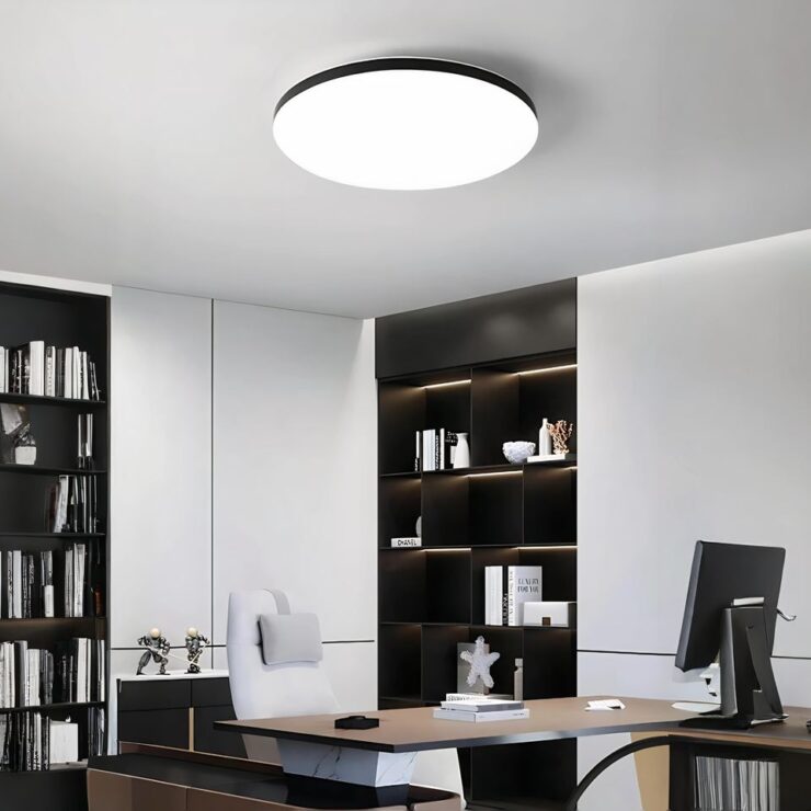 Un lustre plat LED circulaire avec sa base noire. Le cercle entier est lumineux. Il est fixé dans un bureau aux murs blancs avec une étagère murale marron. Chaque est illuminée avec une barre LED à l'intérieur. Des livres et de la déco y sont rangés. Un grand bureau de couleur marron clair avec un écran d'ordinateur, un clavier et des livres sont dessus. Derrière, un fauteuil blanc avec dossier haut. Derrière le fauteuil, un meuble bas marron avec des sujets posés dessus.