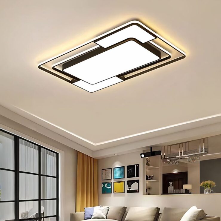 Lustre plat moderne aux lignes géométriques allumé et fixé sur un plafond blanc dans un salon. Sur la gauche une grande fenêtre aux encadrements noirs. Sur la droite, le haut d'un cnaapé beige avec des coussins beiges. Derrière lui, un mur beige avec des tableaux colorés carrés et un comptoir qui sépare d'une autre pièce.
