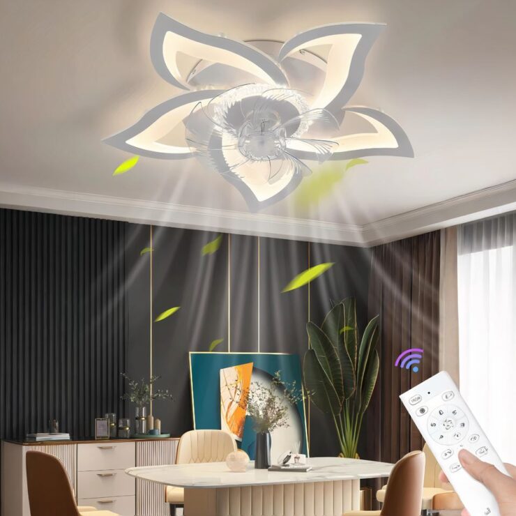 Un lustre ventilateur LED en forme de fleur dans un salon. Le ventilateur à 5 pétales donc les contours intérieurs sont lumineux, l'extérieur est blanc. Il est fixé au-dessus une table de forme ovale beige avec deux chaises marron. Les murs sont marrons foncés, à droite une fenêtre. Dans l'angle se trouve une plante verte. A sa gauche un buffet assorti à la table. Entre les deux, un grand tableau dans les tons vert et beige. En bas à droite de l'imange, une main tenant une télécommande blanche en direction du lustre.