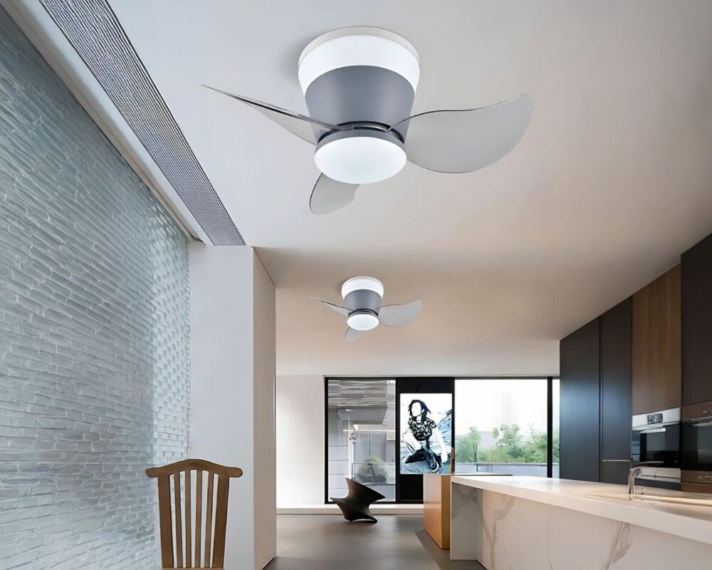 On voir une grande pièce rectangulaire avec sur la gauche, un mur style vitre floutée avec une chaise en bois devant. Sur la droite, un long plan de travail style marbre blanc avec un mur marron foncé sur sa droite. Au plafond, deux lustres ventilateur avec LED intégrées alignés dans la longueur. Ils ont un socle gris et 3 pales transparentes. Au fond de la pièce, un mur beige avec des fenêtres et un tableau au centre.