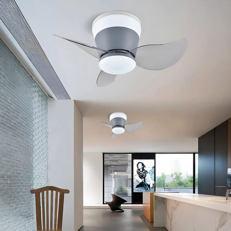 On voir une grande pièce rectangulaire avec sur la gauche, un mur style vitre floutée avec une chaise en bois devant. Sur la droite, un long plan de travail style marbre blanc avec un mur marron foncé sur sa droite. Au plafond, deux lustres ventilateur avec LED intégrées alignés dans la longueur. Ils ont un socle gris et 3 pales transparentes. Au fond de la pièce, un mur beige avec des fenêtres et un tableau au centre.