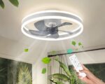 Lustre ventilateur moderne avec éclairage LED en forme d'anneau. L'anneau est blanc avec les LED sur les contours. Le lustre est allumé et fixé sur un plafond blanc.On aperçoit le haut des murs avec sur la gauche un tableau avec de la verdure. Sur la droite un tringle à rideau devant une fenêtre. Une plante verte et devant à droite une main tenant une télécommande blanche en direction du lustre.