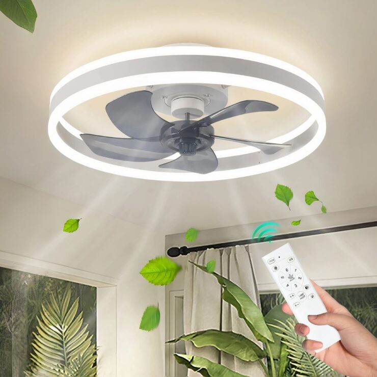 Lustre ventilateur moderne avec éclairage LED en forme d'anneau. L'anneau est blanc avec les LED sur les contours. Le lustre est allumé et fixé sur un plafond blanc.On aperçoit le haut des murs avec sur la gauche un tableau avec de la verdure. Sur la droite un tringle à rideau devant une fenêtre. Une plante verte et devant à droite une main tenant une télécommande blanche en direction du lustre.