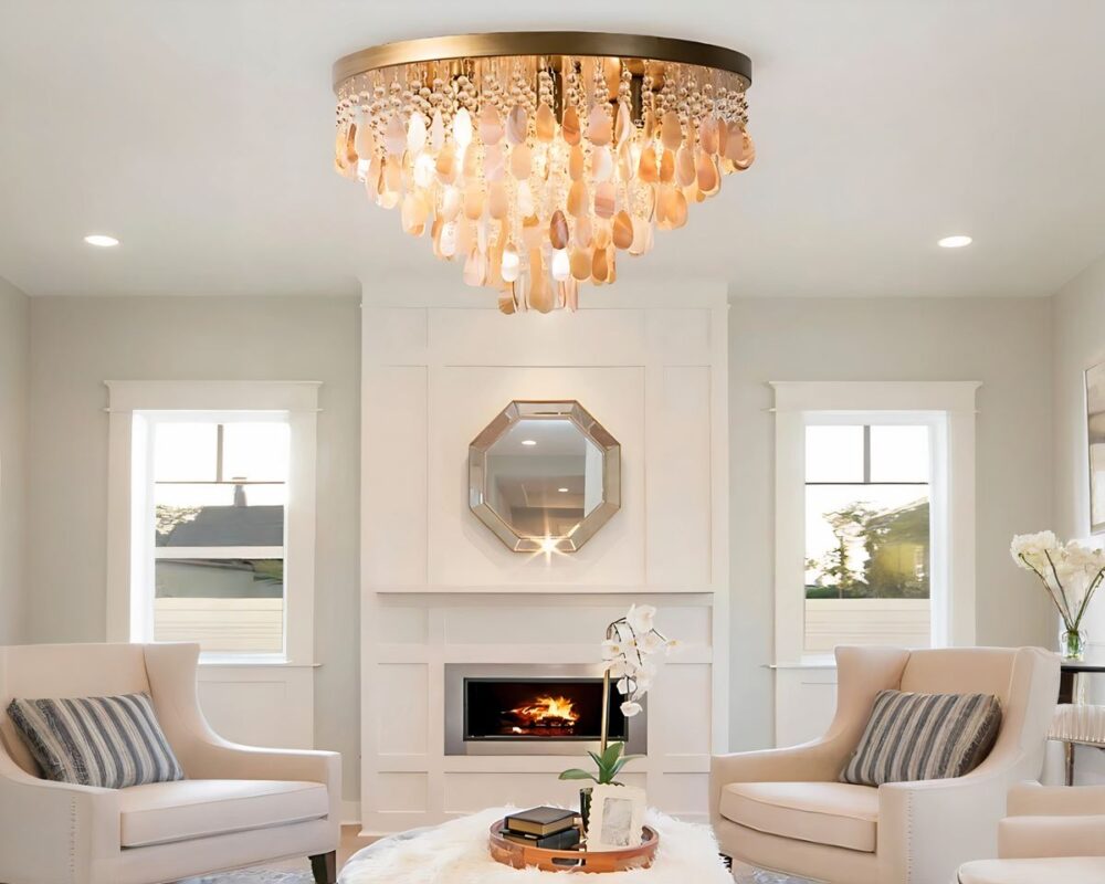 Lustre à pampilles en coquillage beige et style cristal. Le lustre est de style plafonnier rond avec une base dorée fixée au plafond. Les pampilles sont suspendues sur plusieurs niveaux. Il est allumé dans un salon aux murs blancs, au-dessus d'une table basse blanche avec une plat rond et des décos dessus dont une orchidée. De chaque côté de la table un fauteuil beige et un coussin à rayures blanches et bleues. Au fond des murs blancs avec au centre, un insert de cheminée, un miroir au-dessus et de chaque côté une grande fenêtre.