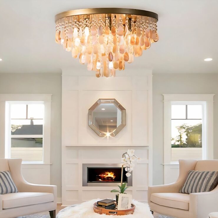 Lustre à pampilles en coquillage beige et style cristal. Le lustre est de style plafonnier rond avec une base dorée fixée au plafond. Les pampilles sont suspendues sur plusieurs niveaux. Il est allumé dans un salon aux murs blancs, au-dessus d'une table basse blanche avec une plat rond et des décos dessus dont une orchidée. De chaque côté de la table un fauteuil beige et un coussin à rayures blanches et bleues. Au fond des murs blancs avec au centre, un insert de cheminée, un miroir au-dessus et de chaque côté une grande fenêtre.