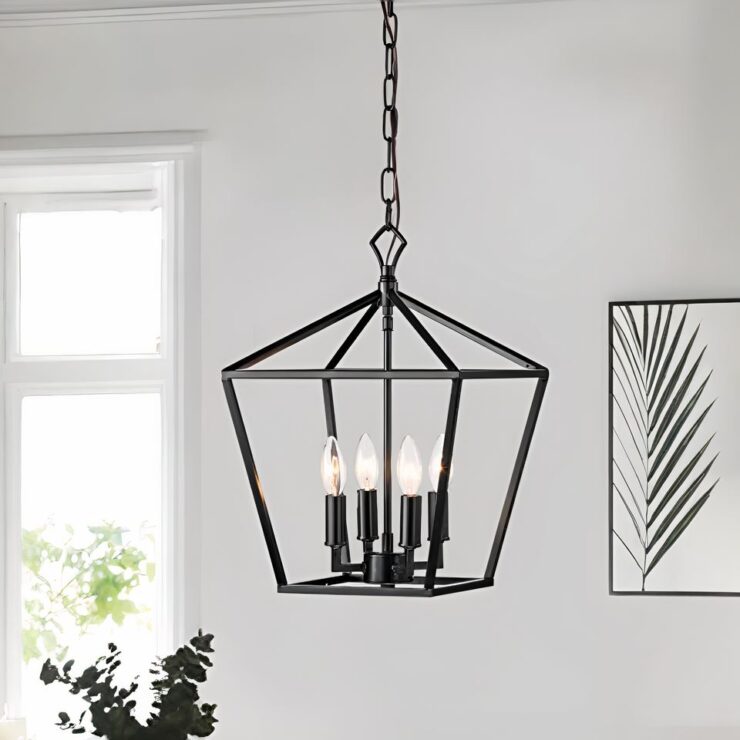 Un lustre cage style américain de couleur noir en métal avec 4 ampoules style chandeliers suspendu par une chaîne en métal. Les ampoules sont allumées. En fond, un mur blanc, avec un tableau sur la droite et une fenêtre à gauche.