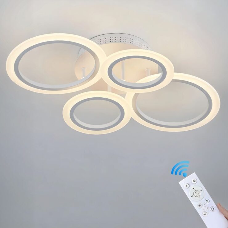 Lustre plafonnier avec 4 cercles lumineux de tailles différentes fixés sur un socle blanc rond. Les anneaux sont allumés. En bas à droite une télécommande blanche avec le symbole en bleu du contrôle en Bluetooth.