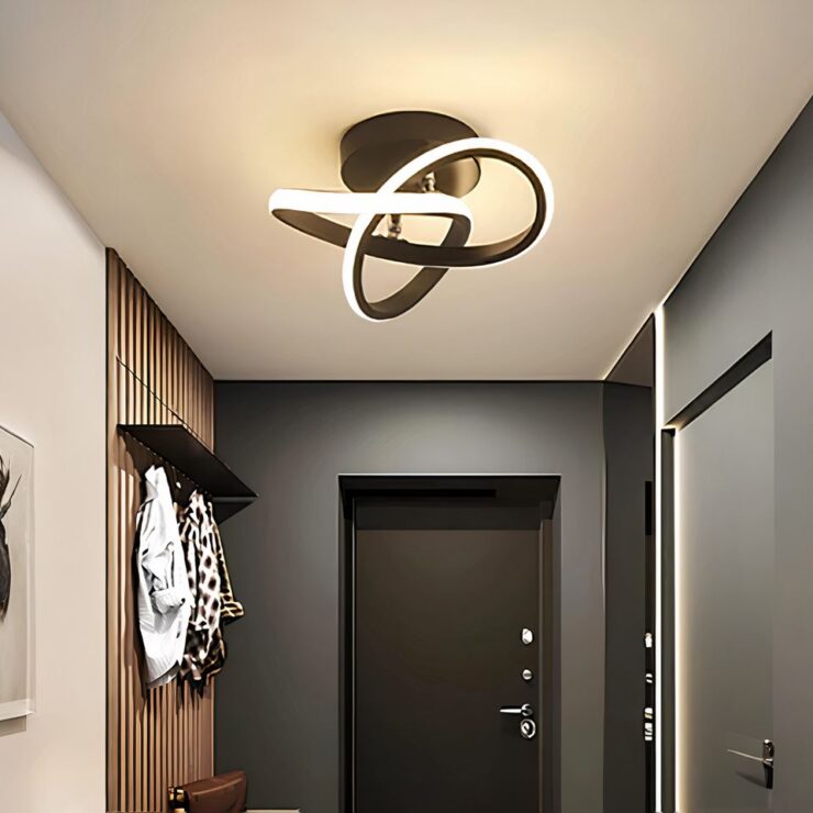 Lustre plafonnier moderne aux courbes entrelacées de couleur noir fixé sur un plafond blanc dans un couloir d'entrée. Le mur de droite est gris, celui d'en face aussi avec une porte d'entrée noire. Le mur de gauche est blanc avec une partie marron où est fixée une étagère porte-manteau avec une veste blanche et une à motifs blancs et noirs. Le lustre est allumé et diffuse une lumière blanc chaud avec ses LED le long des courbes.