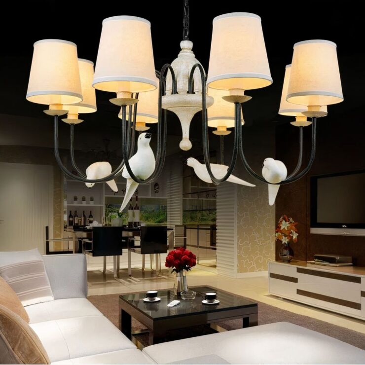 Lustre avec abat-jour romantique et oiseaux perchés, dans sa variante huit abat-jour, suspendu dans un salon, au-dessus d'un canapé blanc et en bois. Le lustre est allumé.