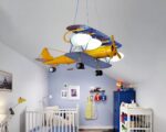 Lustre bébé avion rétro allumé est suspenbdu dans une chambre de bébé. On aperçoit un lit à barreaux ainsi que la table à langer, et le mur du fond est bleu.