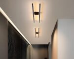 L'image montre 2 lustres minimalistes avec deux barres LED parallèles, chacun de couleur noire, l'un derrière l'autre fixés sur le plafond d'un couloir. Ils diffusent une lumière blanc chaud. Sur la gauche, on aperçoit un mur marron foncé, style placard.