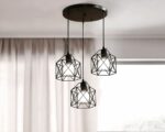 Lustre 3 suspensions en forme de cage en métal noir style industriel avec une ampoule dans chaque cage. Les 3 cages sont suspendues par un câble noir fixé sur un support rond noir au plafond. On aperçoit une fenêtre avec un voilage beige en arrière plan.