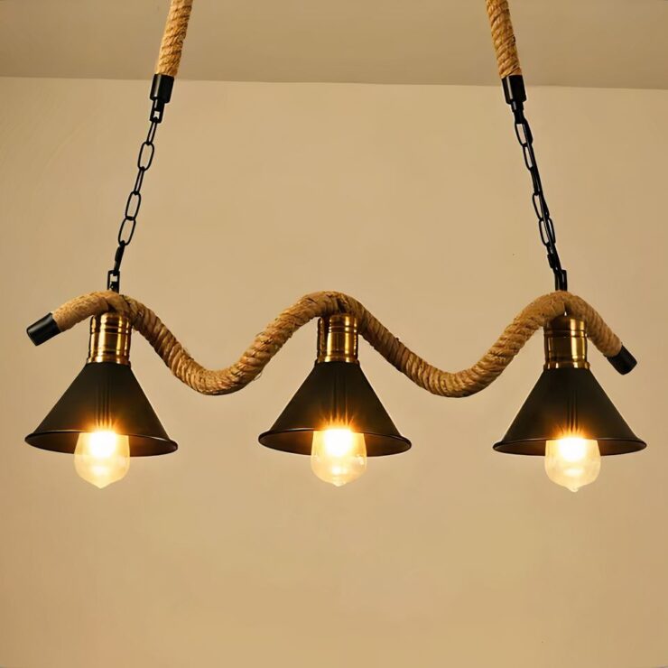 On voit un lustre 3 suspensions industrielles avec corde sur fond marron. Le lustre est composé de trois abats-jour en métal noir de forme conique avec une ampoule allumée. Ses abats-jour sont fixés sur une corde ondulée qui elle-même est suspendue par deux chaînes avec le haut en corde.