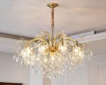 On voit un lustre chic avec pampilles en cristal. Il est composé d'un socle rond foré fixé sur un plafond blanc avec une chaîne où sont suspendues plusieurs branches dorées. Sue chaque branches, plusieurs pampilles en cristal transparent. Les ampoules dissimulées entre les pampilles sont allumées. Les murs de la pièce sont de couleur blanc/beige.