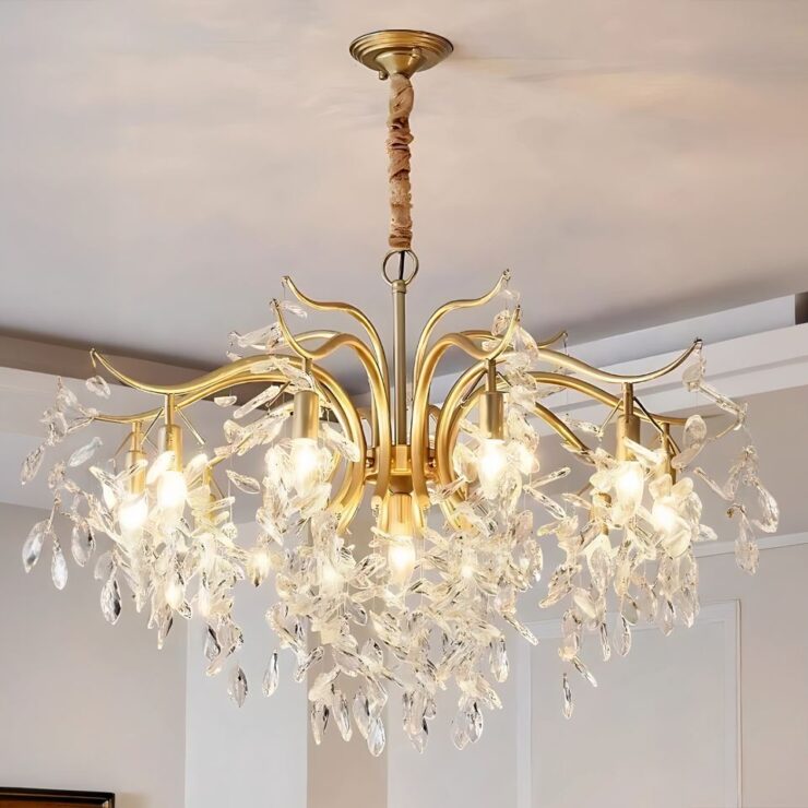 On voit un lustre chic avec pampilles en cristal. Il est composé d'un socle rond foré fixé sur un plafond blanc avec une chaîne où sont suspendues plusieurs branches dorées. Sue chaque branches, plusieurs pampilles en cristal transparent. Les ampoules dissimulées entre les pampilles sont allumées. Les murs de la pièce sont de couleur blanc/beige.