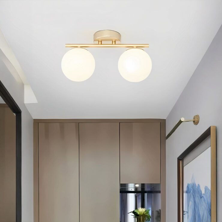 Lustre d'entrée à deux boules lumineuses tenues sur une barre dorée. Le lustre plafonnier est fixé dans un couloir d'entrée avec à droite un mur avec un tableau. Au fond un mur style bois moderne et une petite étagère intégrée. Sur la gauche, une porte d'entrée marron avec un encadrement noir.