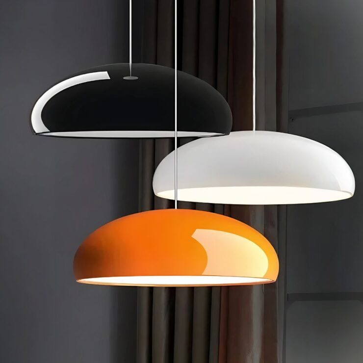 On voit trois lustres d'entrée colorés au design moderne. Ils sont suspendus à différentes hauteurs de couleur noir, blanc et orange. Ils sont allumés. Ils sont en forme de demi-ovale. En fond un mur gris foncé avec un rideau marron.