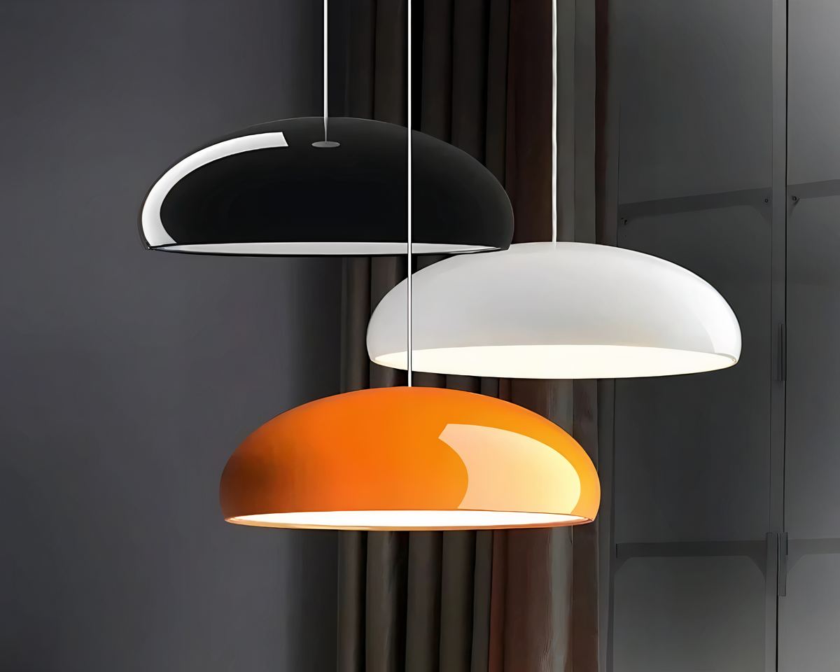 On voit trois lustres d'entrée colorés au design moderne. Ils sont suspendus à différentes hauteurs de couleur noir, blanc et orange. Ils sont allumés. Ils sont en forme de demi-ovale. En fond un mur gris foncé avec un rideau marron.