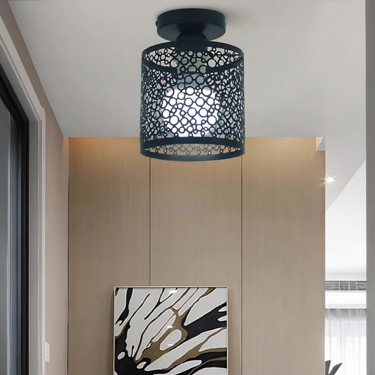 Lustre d'entrée plafonnier avec abat-jour élégant noir au design ajouré fixé sur un plafond blanc. Il est composé d'un abat-jour cylindrique en métal ajouré laissant voir l'ampoule allumée. En face, un mur beige avec un tableau noir et blanc. Sur la gauche, on aperçoit une porte foncée.