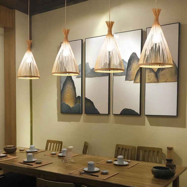 On se trouve dans une salle à manger avec une grande table rectangulaire en bois avec des tasses blanches dessus et des chaises en bois autour. Le mur derrière est beige avec 4 tableaux blancs. Au dessus de la table, 4 lustres japonais nautrels en bambou identiques de couleur naturelle beige et allumés.