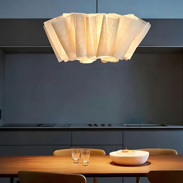 Un lustre original avec abat-jour en lin plissé dont la lumière est allumée. Il est fixé au dessus d'une table en bois avec des chaises assorties autour. Sur la table deux verres et un plat blanc. En fond un plan de travail de cuisine gris foncé avec le mur assorti.