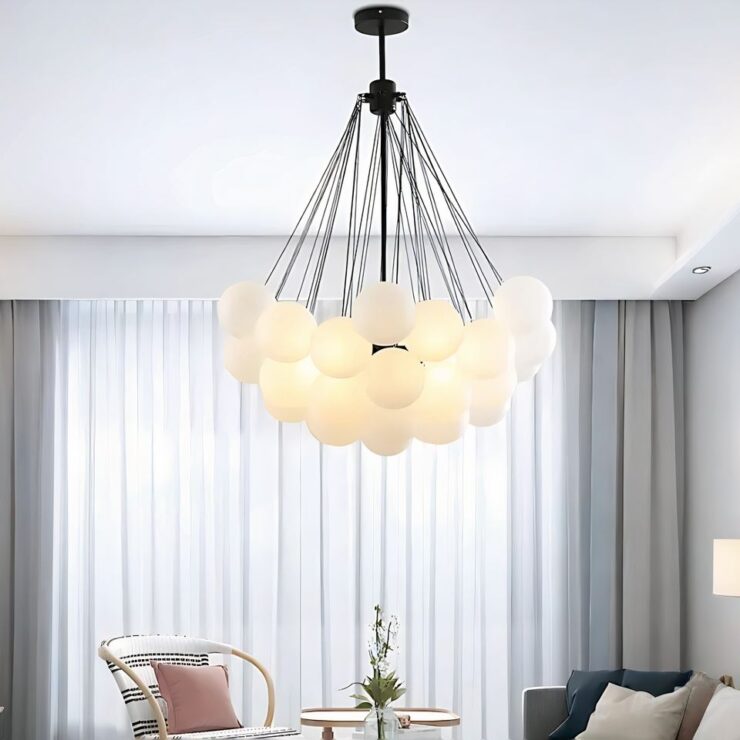 Lustre original avec grappe de boules lumineuses fixé au centre d'un salon. La structure est composée un socle noir noir duquel sont suspendues les boules lumineuses en verre givré par des chaines en métal noir. Au fond on voit une grande fenêtre qui prend toute la longueur du mur, avec des voilages blancs et des rideaux gris sur les côtés. Devant, un fauteuil blanc à rayures avec des accoudoirs en bois. A droite, un canapé et une table basse.