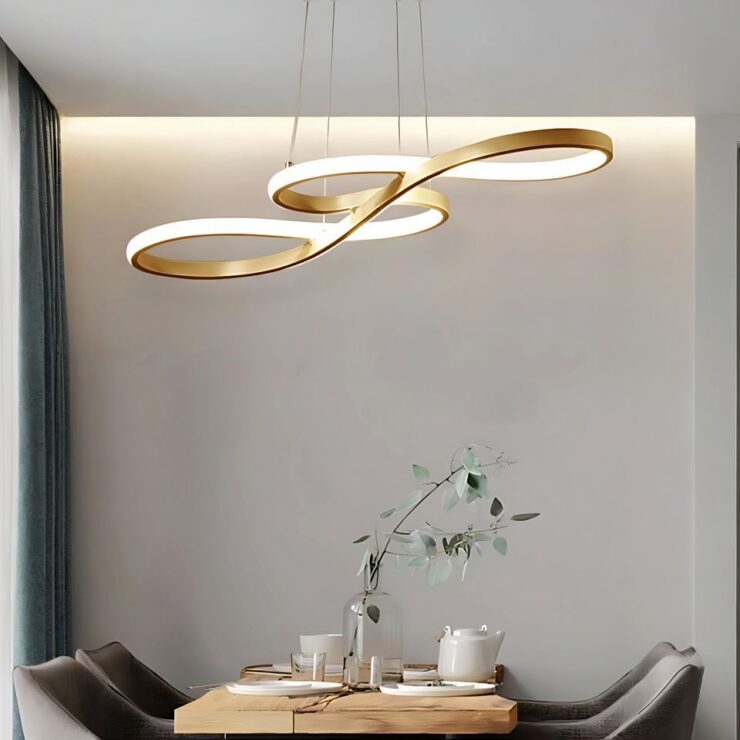 On voit un lustre salle à manger LED aux lignes fluides de couleur doré avec le contour lumineux. Il ressemble à une clé de sol. Suspendu par des câbles fins. Il est fixé au-dessus une table rectangle beige avec des fauteuils gris de chaque côté. Sur la table de la vaisselle blanche. Les murs sont gris.