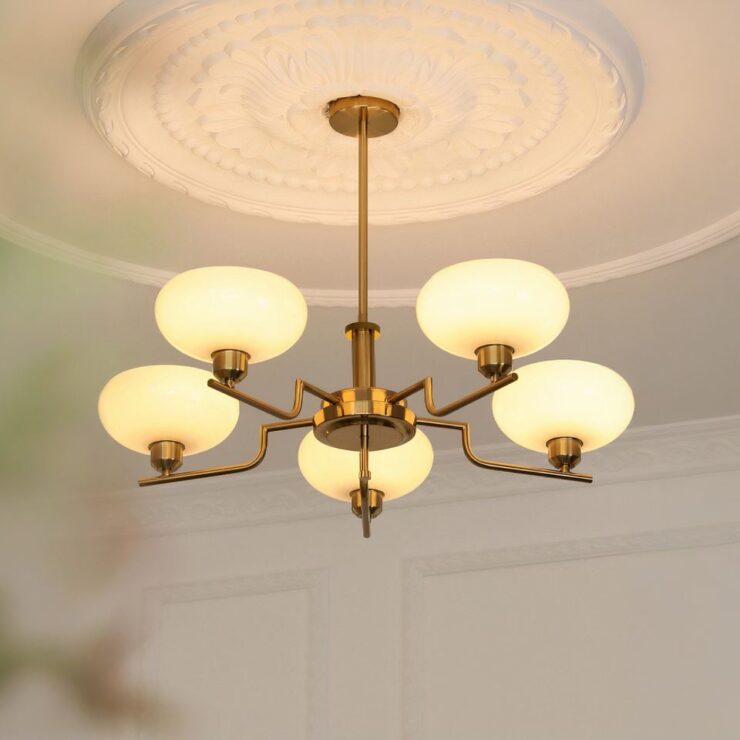 On voit un lustre salle à manger au style rétro. Le plafond est blanc avec un cercle en relief. Le lustre est composé d'un socle rond doré avec une tige dorée qui descend, reliée à 5 branches qui ont chacune un globe ovale en verre. Chaque ampoule est allumée d'une lumière blanc chaud. Les murs autour sont blancs.