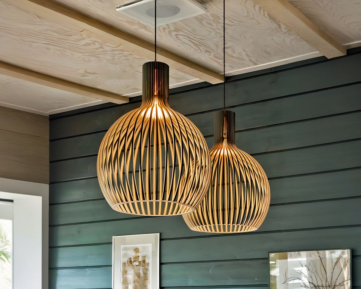 On aperçoit une pièce avec un mur en lattes de bois peintes en vert, le plafond est bois beige. Sur le mur vert, deux petites cadres. Sont suspendus au plafond, deux lustres salle à manger en bois ajouré de forme ronde laissant passer la lumière à travers les lamelles de bois.