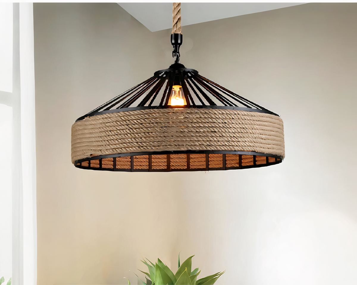 Lustre salle à manger style industriel avec corde suspendu et allumé. Il est suspendu par une corde. L'abat-jour est en métal noir de forme circulaire avec de la corde sur le contour. L'ampoule est allumée. Les murs sont beiges. Le haut d'une plante verte est visible sur le bas de l'image.