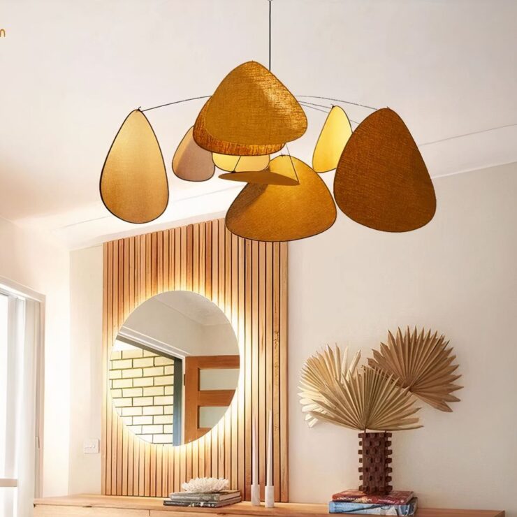 Lustre salon avec facettes en tissu marron fixé dans une pièce aux murs blancs. Le lustre est allumé. Il est composé d'une tige noire suspendue au plafond qui relie plusieurs tiges horizontales sur lesquelles sont suspendues des facettes de forme triangulaire en tissu. Derrière on aperçoit un meuble en bois avec un miroir rond. De la déco y est posée.