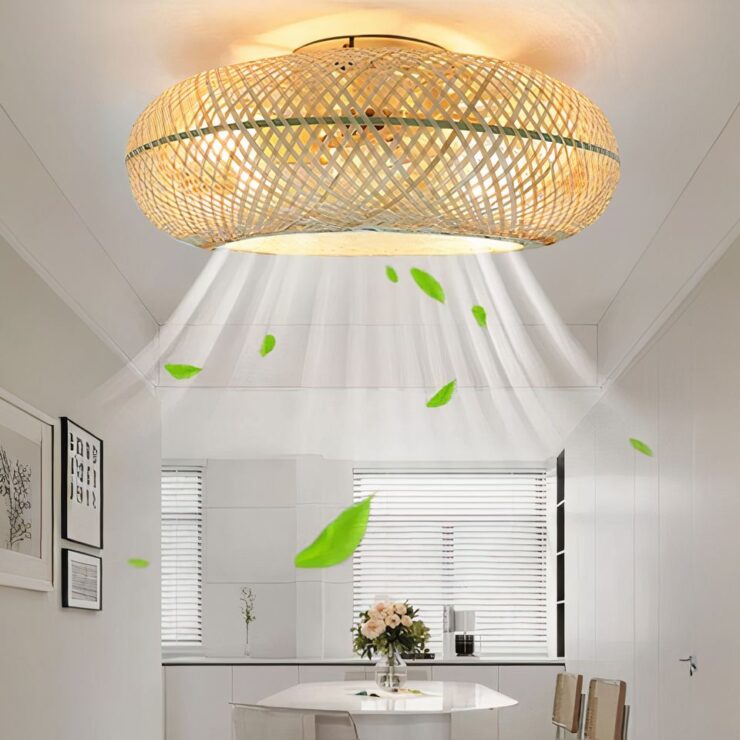 On voit un lustre salon en rotin avec ventilateur intégré. La lumière est allumée. Il est fixé sur un plafond blanc, dans une pièce étroite aux murs blancs. Sur le mur de gauche, trois cadres. Au fond une table ovale blanche, deux chaises à droite de la table. Au fond de la pièce, un plan de travail avec des fenêtres.