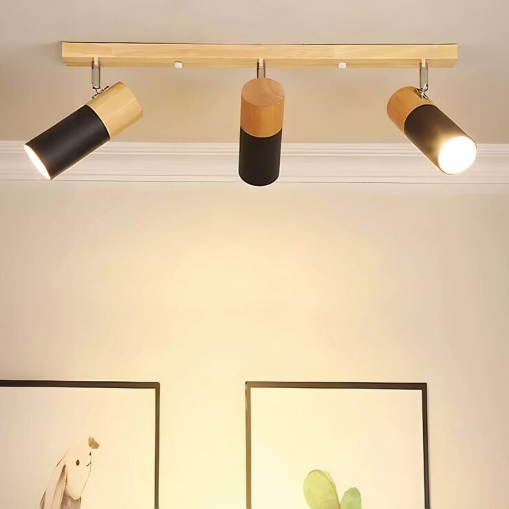 Lustre spot style nordique en bois fixé sur un plafond beige. Le mur à l'arrière est beige avec deux tableaux aux encadrements noirs avec un lapin dessiné à gauche et un cactus à droite. Le lustre plafonnier est composé d'une barre en bois clair sur laquelle sont fixées 3 spots cylindriques allumés. La partie haute du spot est en bois et la partie basse est noire. Le spot de gauche est orienté vers la gauche, le spot du milieu est arrière vers le mur et le spot de droite est orienté vers la droite.
