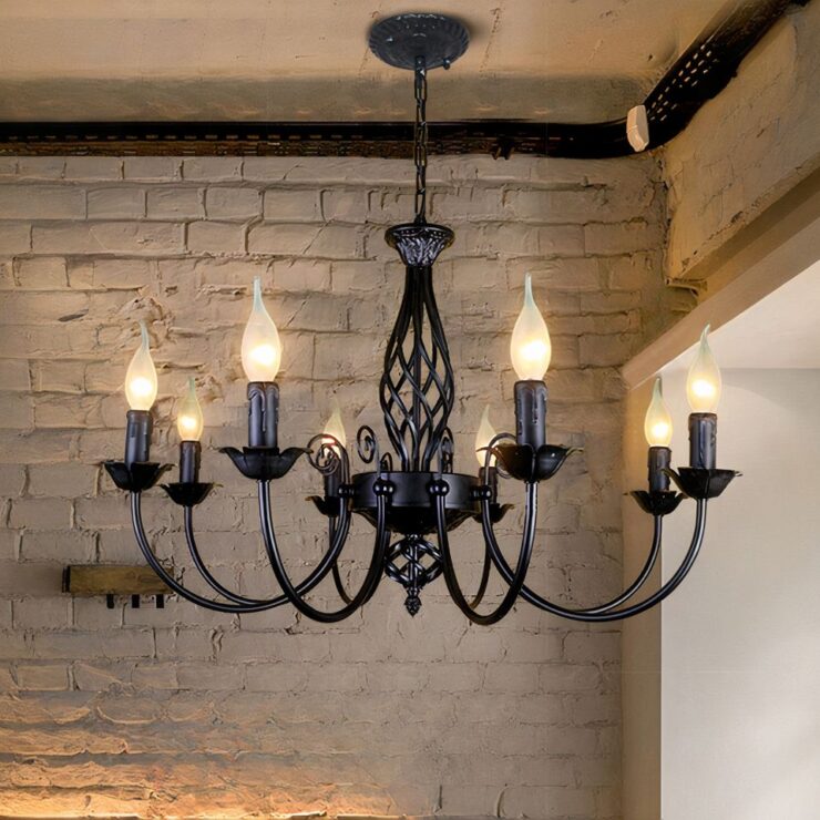 Un lustre baroque en fer forgé avec bougies LED possédant huit bras courbés, chacun équipé d'ampoules LED en forme de bougie. Il est suspendu par une chaîne dans une pièce avec un mur en briques apparentes.
