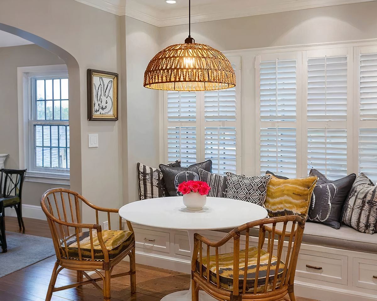 Une petite salle à manger cosy avec une table ronde blanche, entourée de chaises en rotin. Au-dessus de la table, un lustre bohème en chanvre cordé est suspendu, il éclaire la pièce avec une lumière douce et chaleureuse. Un coin banquette avec des coussins colorés est disposé sous une grande fenêtre à volets blancs.