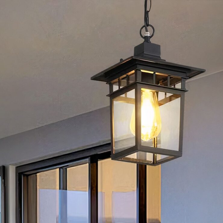 Un lustre extérieur moderne en métal noir est suspendu au plafond d’une terrasse extérieure. Il est de forme rectangulaire, avec des panneaux en verre transparent entourant une grande ampoule. Il diffuse une lumière douce.
