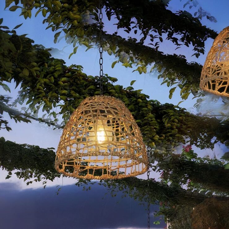 Un lustre extérieur solaire de style bohème en rotin est suspendu à une chaîne, accroché à une branche d’arbre dans un jardin. Il diffuse une lumière douce dans l’obscurité, créant une ambiance chaleureuse et accueillante.