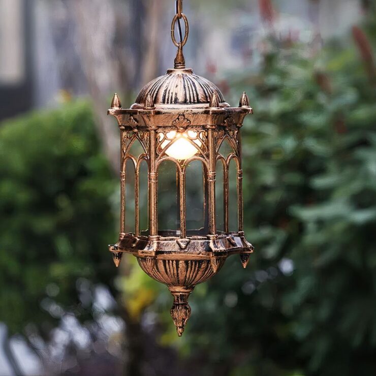 Un lustre extérieur de style gothique en métal doré est suspendu par une chaîne métallique dans un cadre extérieur verdoyant. Ce lustre est cylindrique avec une lumière centrale à l'intérieur, protégée par des panneaux en verre.