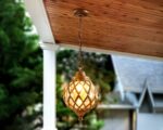 Un lustre extérieur pour terrasse style bohème chic est suspendu sous une pergola. Il est en métal doré avec un globe en verre et diffuse une lumière douce. Ce luminaire ajoute une ambiance chaleureuse et élégante à l'espace extérieur.